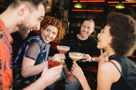 Despreocupada pelirroja sosteniendo cóctel y mirando a amigos interracial hablando en el bar 