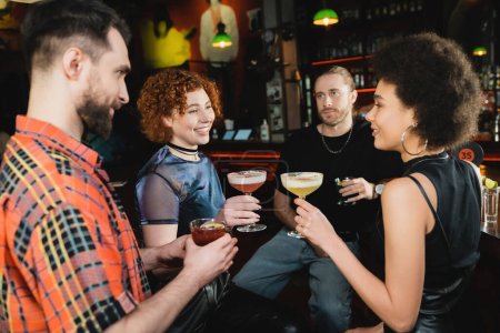 Mujeres multiétnicas positivas celebrando cócteles y hablando cerca de amigos en el bar 