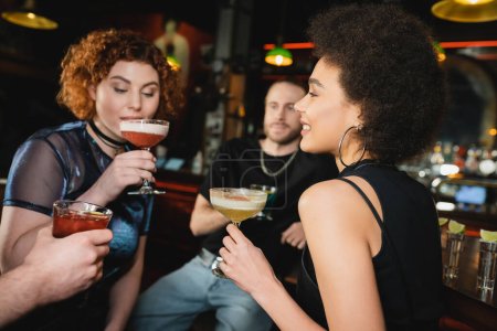 Positive curly african american woman holding pisco sour cocktail near friends in bar 