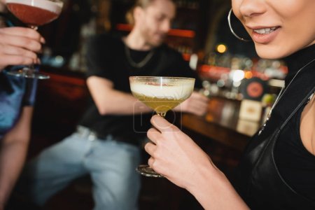 Foto de Vista recortada de la sonriente mujer afroamericana sosteniendo cóctel de espuma mientras pasa tiempo con amigos en el bar - Imagen libre de derechos
