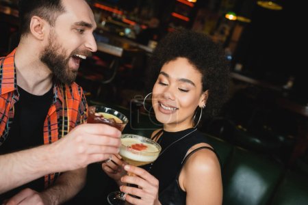 Excited man holding negroni cocktail near curly african american friend in bar 