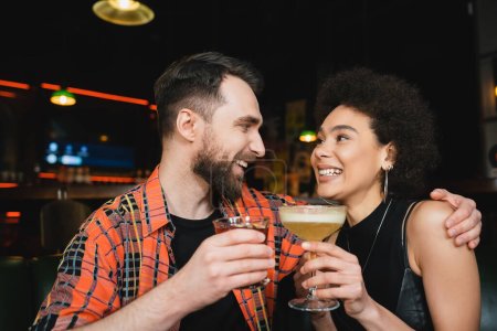 Hombre alegre celebración de cóctel y abrazo rizado amigo afroamericano en el bar 