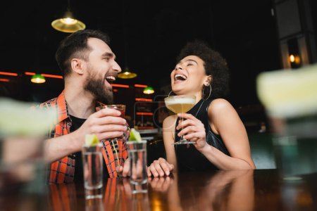 Laughing african american woman holding cocktail near friend and tequila shots in bar 