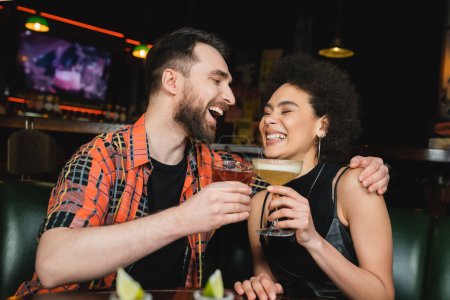 Lachender Mann hält Negnoni-Cocktail in der Hand und umarmt afrikanisch-amerikanischen Freund neben verschwommenem Tequila in Bar 