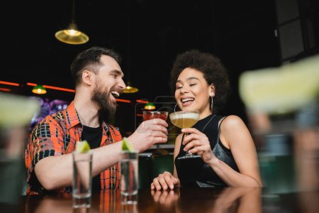 Overjoyed interracial friends holding cocktails near tequila shots on stand in bar 