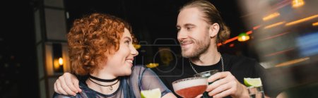 Smiling bearded man hugging redhead friend with cocktail in bar, banner 