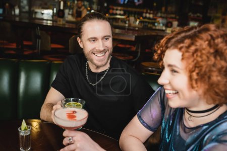 Cheerful man holding blue lagoon cocktail near blurred redhead friend in bar 