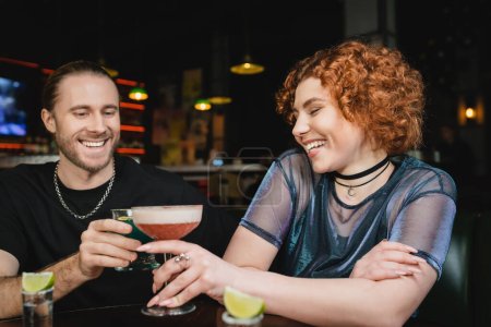 Smiling redhead woman clinking cocktail with bearded friend in bar 