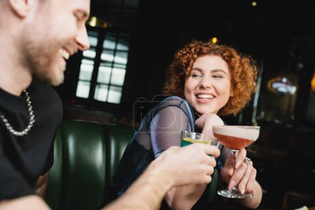 Positive redhead woman clinking clover club cocktail with blurred friend in bar 