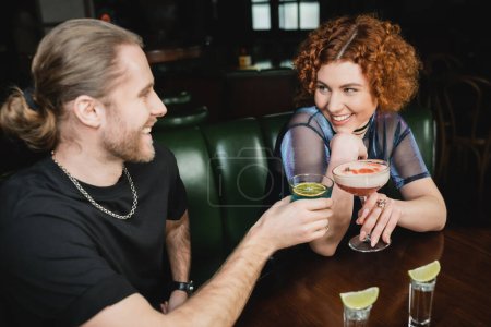 Fröhliche rothaarige Frau hält Kleeblatt-Cocktail neben Freundin und steht in Bar 