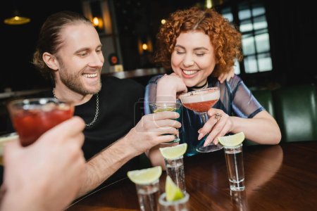 Amigos sonrientes tintinean diferentes cócteles cerca de tiros de tequila con lima en el bar 