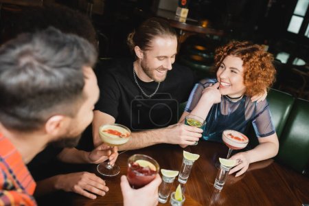 Smiling man holding blue lagoon cocktail near multiethnic friends spending time in bar 