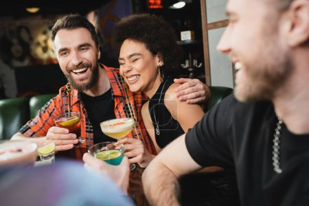 Fröhlicher Mann umarmt lockigen afrikanisch-amerikanischen Freund mit Cocktail in Bar 