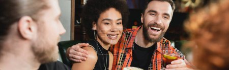 Smiling man holding negroni cocktail and hugging african american friend in bar in evening, banner 