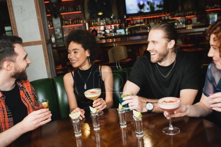 Hombre barbudo positivo hablando con amigos multiétnicos con cócteles cerca de tequila en el bar 