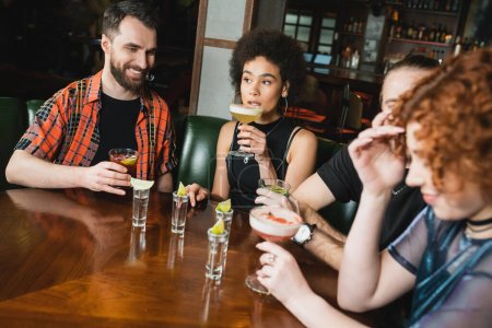 Amigos multiétnicos con cócteles y tragos de tequila pasando tiempo en el bar por la noche 