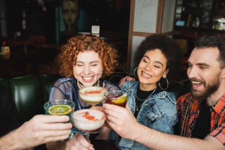 positive interracial friends smiling and toasting with different cocktails in bar 