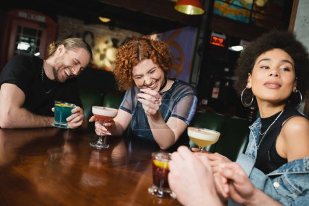 happy interracial friends laughing and holding different cold cocktails in bar 