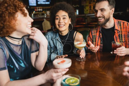 Jeune femme afro-américaine frisée tenant un cocktail près d'amis gais dans un bar 