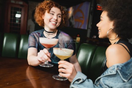 Smiling interracial girlfriends holding blurred cocktails in bar 