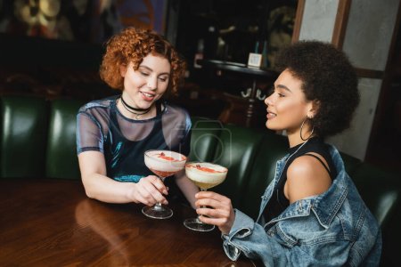 Redhead woman holding clover club cocktail and talking to african american friend in bar  