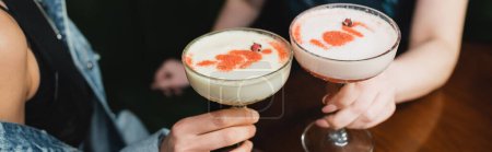Cropped view of multiethnic girlfriends holding cold foam cocktails in bar in evening, banner 