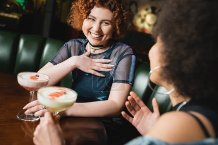 Carefree woman holding clover club cocktail near blurred african american girlfriend in bar 