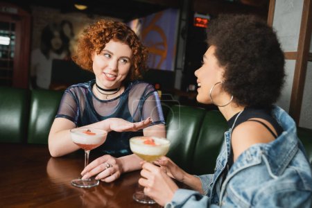 Smiling red haired woman talking to african american girlfriend near cocktails in bar 