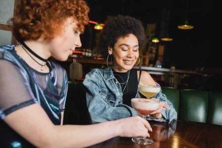 Smiling african american woman looking at pisco sour cocktail near friend in bar 
