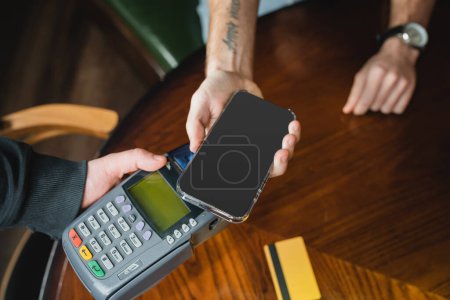 Vista recortada del hombre que paga con teléfono inteligente cerca de camarero con terminal de pago en el bar 