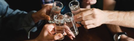 Cropped view of multiethnic friends clinking tequila shots in bar in evening, banner 