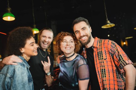 Téléchargez les photos : Homme excité montrant geste rock près d'amis multiethniques passer du temps dans le bar en soirée - en image libre de droit