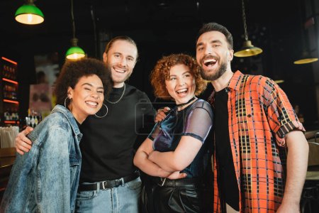 Cheerful redhead woman crossing arms while multiethnic friends looking at camera in bar 