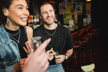 Smiling bearded man holding tequila shot near multiethnic friends in bar 