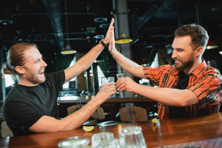 Carefree men holding tequila shots and giving high five near lime in bar at night 