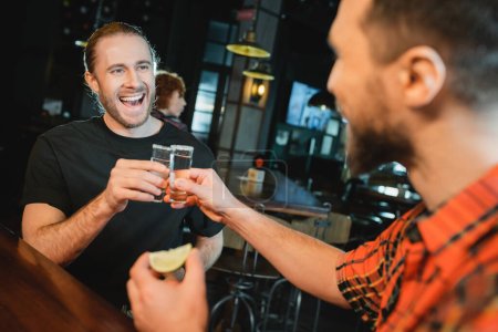 Photo for Smiling bearded man clinking tequila shots with blurred friend in bar - Royalty Free Image