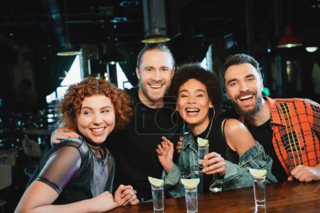 Cheerful interracial friends looking at camera near glasses with tequila in bar 
