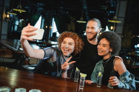 Téléchargez les photos : Ami multiethnique joyeux prenant selfie sur smartphone près de la tequila dans le bar - en image libre de droit