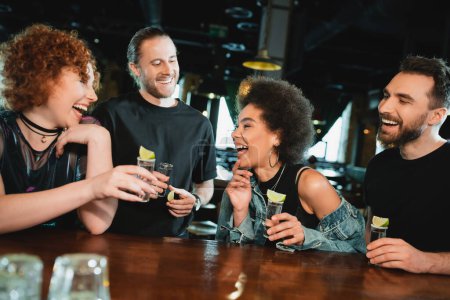 Carefree interracial people holding tequila shots near stand in bar 