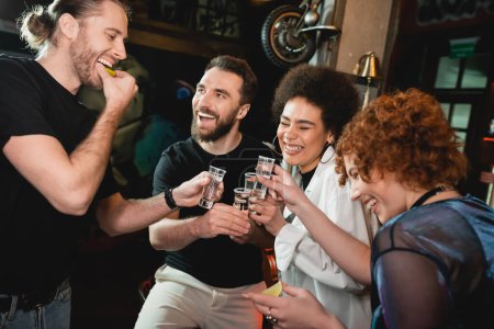 Excited interracial friends holding tequila shots and fresh lime in bar 