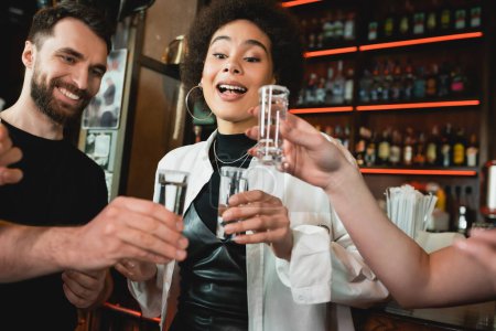 Excited african american woman holding tequila shot with salt near friends in bar 