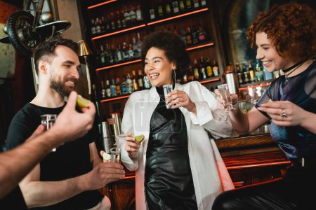 Multiethnic friends holding tequila and fresh lime near stand in bar 