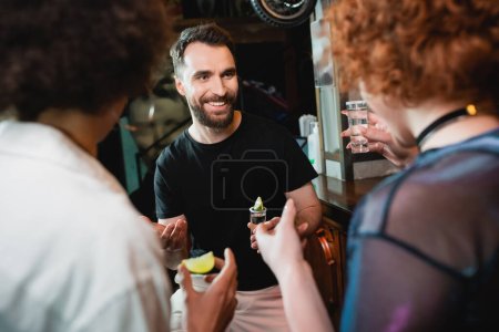 Smiling bearded man talking to blurred interracial friends with tequila in bar 