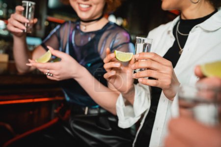 Cropped view of smiling multiethnic women holding tequila and lime in bar 