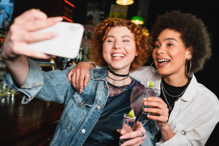 Positive Frau macht Selfie mit afrikanisch-amerikanischer Freundin und hält Tequila in Bar 