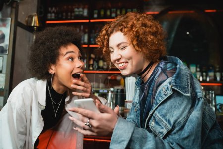 Téléchargez les photos : Femme rousse excitée utilisant un smartphone près d'un ami afro-américain dans un bar - en image libre de droit