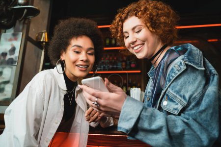 Amigos multiétnicos positivos utilizando el teléfono inteligente cerca de stand en el bar por la noche 