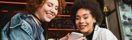 Positive interracial young women using cellphone in bar, banner 