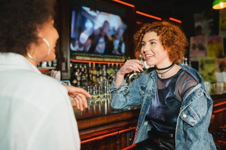 Young redhead woman talking to blurred african american friend near stand in bar 