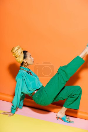 Téléchargez les photos : Vue latérale de la femme afro-américaine à la mode en foulard et vêtements lumineux sur fond coloré - en image libre de droit